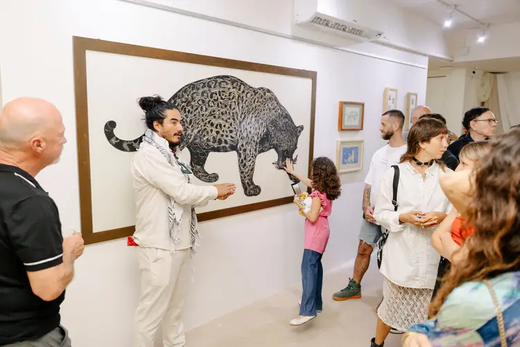 Man in cream suit gestures toward framed drawing of a leopard while visitors gather around in an art gallery space