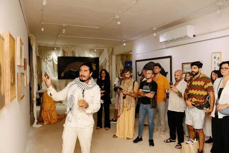 A man in a white outfit gestures while speaking to a group of visitors gathered inside a gallery space with artwork on the walls