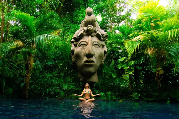 Person in meditation pose floating in pool beneath large stone face sculpture surrounded by tropical vegetation