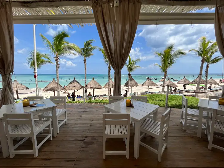 Open-air beachfront restaurant with white tables and chairs overlooking palm trees, thatched umbrellas, and turquoise water under blue sky