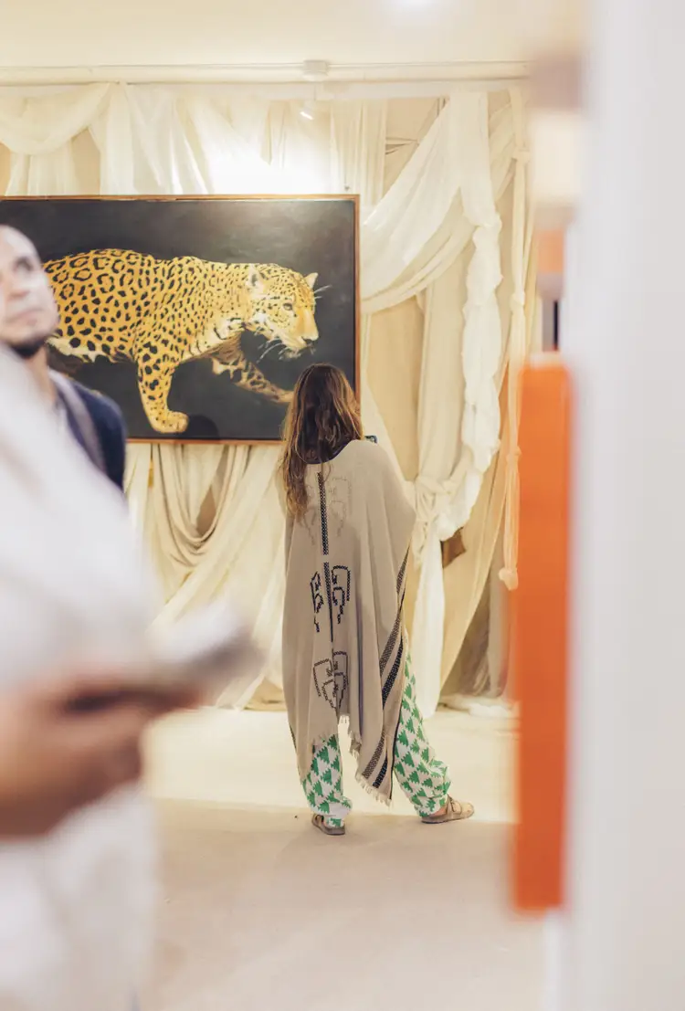 Woman viewing a framed painting of a spotted leopard on a black background in an art gallery with cream-colored draped walls