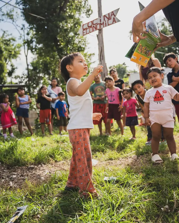 A young girl reaches upward while holding a book in an outdoor gathering of children beneath trees and a wooden sign