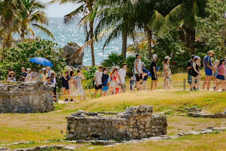 A group of tourists walks through a grassy area with stone ruins, palm trees, and ocean views in the background A group of tourists walks through a grassy area with stone ruins, palm trees, and ocean views in the background