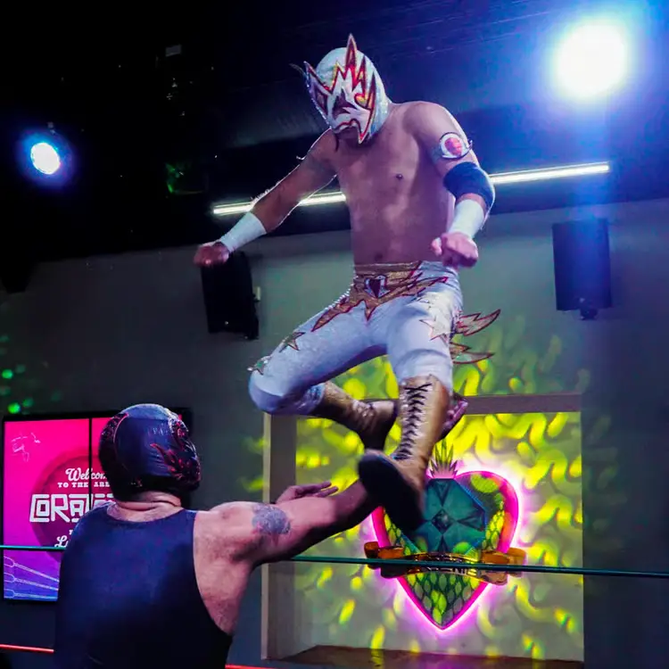 A shirtless wrestler wearing a red and white mask leaps above an opponent during a professional wrestling match in an indoor arena lit by blue stage lights
