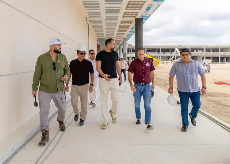 A group of men in casual clothing walk along a concrete pathway inside a large commercial building under construction, with solar panels visible on the roof abo A group of men in casual clothing walk along a concrete pathway inside a large commercial building under construction, with solar panels visible on the roof abo