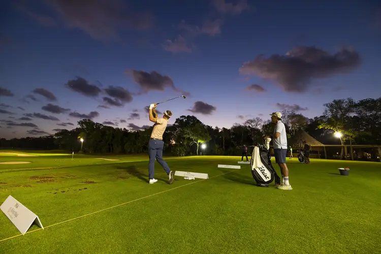 Golfers practice on a floodlit driving range at dusk with trees lining the course in the background