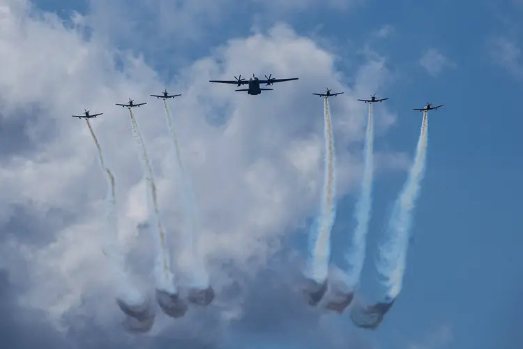 Military transport planes fly in formation while releasing smoke trails against a cloudy blue sky