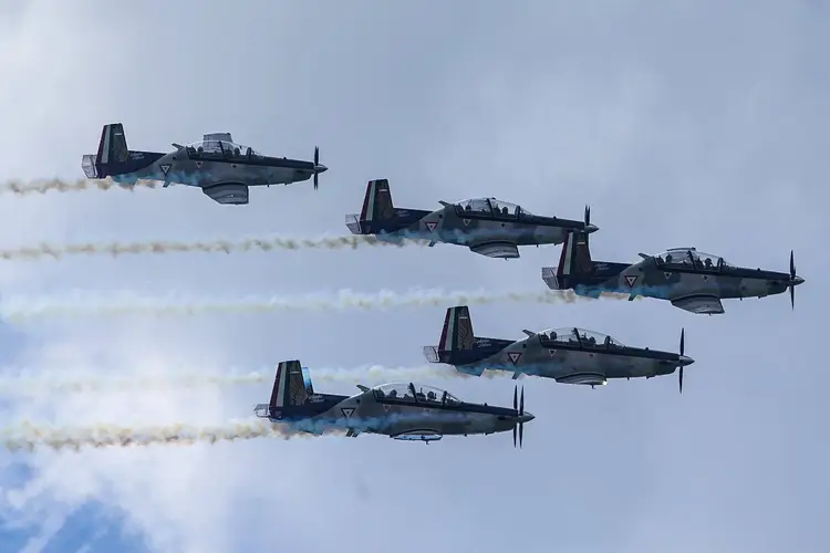 Five military trainer aircraft fly in formation while releasing smoke trails against a blue sky