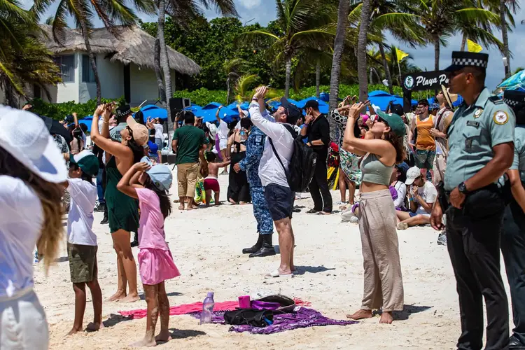 A large crowd gathers on a sandy beach near thatched-roof buildings and palm trees, with uniformed officials present among visitors and beachgoers under blue um