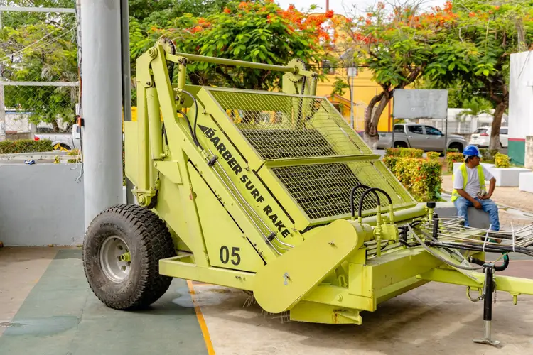 A bright yellow sargassum collection machine sits parked on a paved area with a worker nearby, surrounded by tropical vegetation and buildings A bright yellow sargassum collection machine sits parked on a paved area with a worker nearby, surrounded by tropical vegetation and buildings