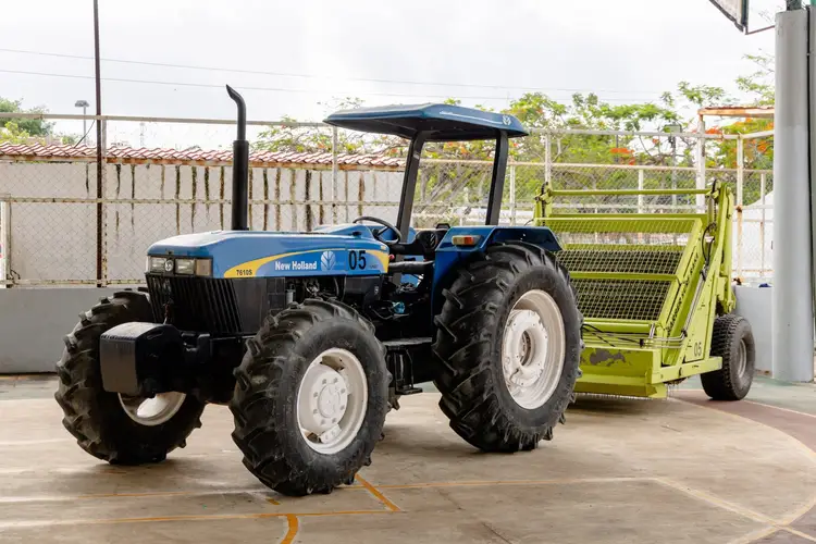 Blue and black tractor with canopy attached to a yellow-green collection trailer on a concrete surface in an industrial yard Blue and black tractor with canopy attached to a yellow-green collection trailer on a concrete surface in an industrial yard