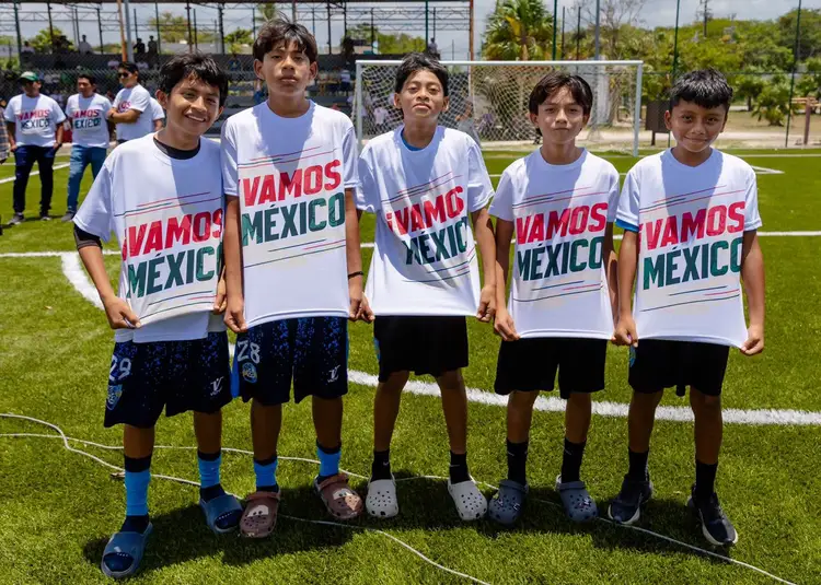 Five young soccer players stand on a grass field wearing matching white shirts with text, with a sports complex and other people visible in the background Five young soccer players stand on a grass field wearing matching white shirts with text, with a sports complex and other people visible in the background