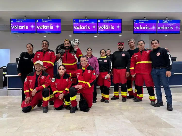 A group of firefighters and rescue workers in red uniforms and protective gear pose together indoors beneath digital signage, with one person holding a dog