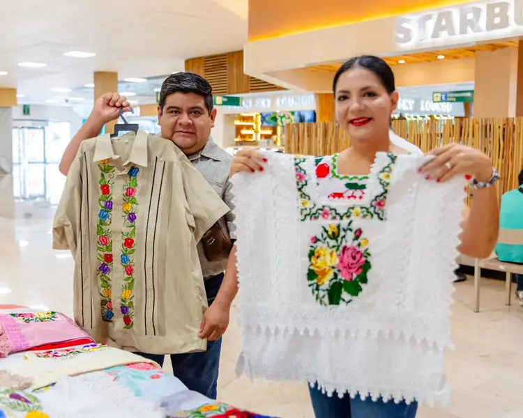 Two people display embroidered clothing with colorful floral designs at a table inside an airport terminal with a Starbucks storefront visible in the background Two people display embroidered clothing with colorful floral designs at a table inside an airport terminal with a Starbucks storefront visible in the background