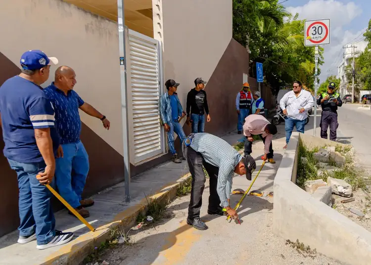 Multiple workers and residents gather on a sidewalk near brown residential buildings while one person uses a measuring tool on the street pavement Multiple workers and residents gather on a sidewalk near brown residential buildings while one person uses a measuring tool on the street pavement