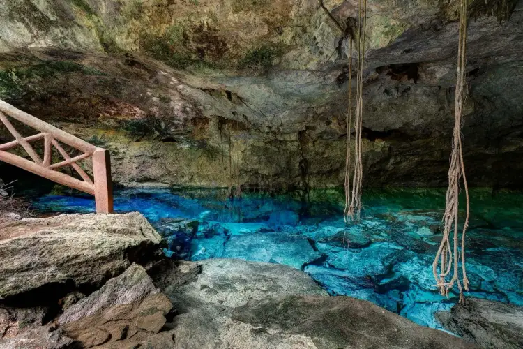 Underground cenote with bright turquoise water, wooden platform, ropes, and limestone cave formations overhead Underground cenote with bright turquoise water, wooden platform, ropes, and limestone cave formations overhead