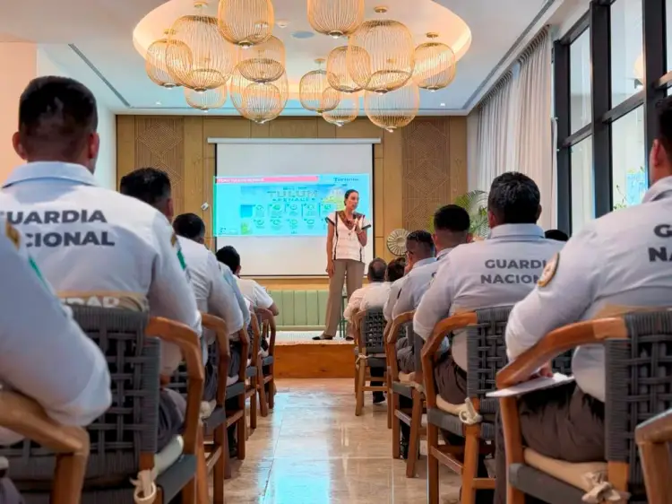 Uniformed officers sit in rows facing a presenter standing before a projection screen in a meeting room with wooden paneling and hanging lights