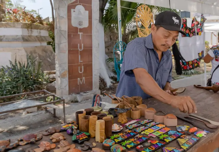 Man in blue shirt and black cap carves wood at a table displaying wooden crafts and colorful painted items in an outdoor market area Man in blue shirt and black cap carves wood at a table displaying wooden crafts and colorful painted items in an outdoor market area