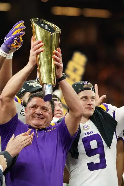 Two people in purple and gold team attire hold a large gold trophy aloft while celebrating on a field