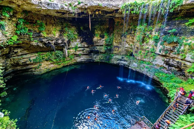 Multiple swimmers and snorkelers in a deep underground cenote surrounded by moss-covered limestone cliffs and a wooden staircase Multiple swimmers and snorkelers in a deep underground cenote surrounded by moss-covered limestone cliffs and a wooden staircase