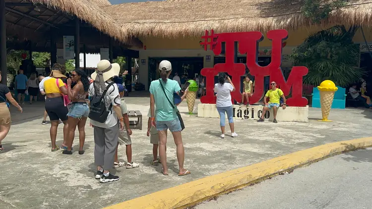 Visitors stand in a plaza near large red letters spelling a word, with thatched-roof structures and tropical vegetation visible behind them Visitors stand in a plaza near large red letters spelling a word, with thatched-roof structures and tropical vegetation visible behind them