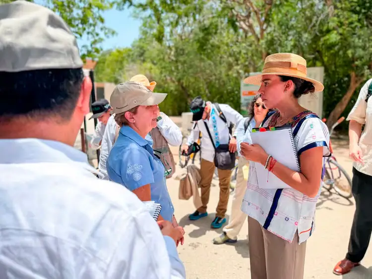 A woman in a tan hat and white shirt speaks to a group gathered outdoors near trees on a sandy area