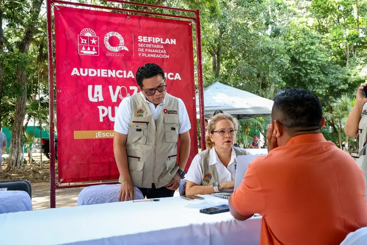 Government officials sit at a table during a public information session in a park, with a red banner behind them and attendees approaching to speak with them Government officials sit at a table during a public information session in a park, with a red banner behind them and attendees approaching to speak with them