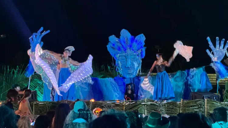 Illuminated blue theatrical figures and performers on stage at a nighttime outdoor event with audience members watching in the foreground