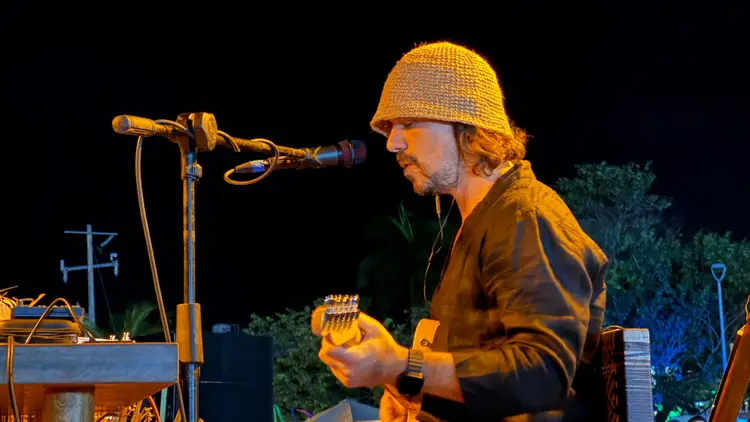 Musician performs at a nighttime outdoor event, singing into a microphone while wearing a knit hat and playing a handheld instrument