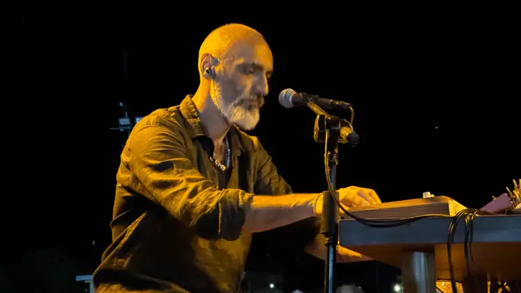 Man with a beard performs at a keyboard while singing into a microphone on a dark stage