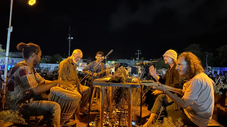Musicians perform with traditional drums and instruments at an outdoor nighttime gathering with spectators seated in the background