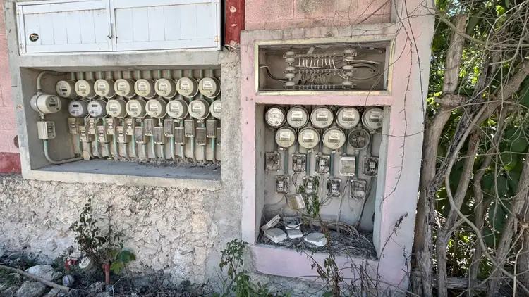 Old electrical meter boxes mounted on a weathered pink building wall with exposed wiring and overgrown vines nearby