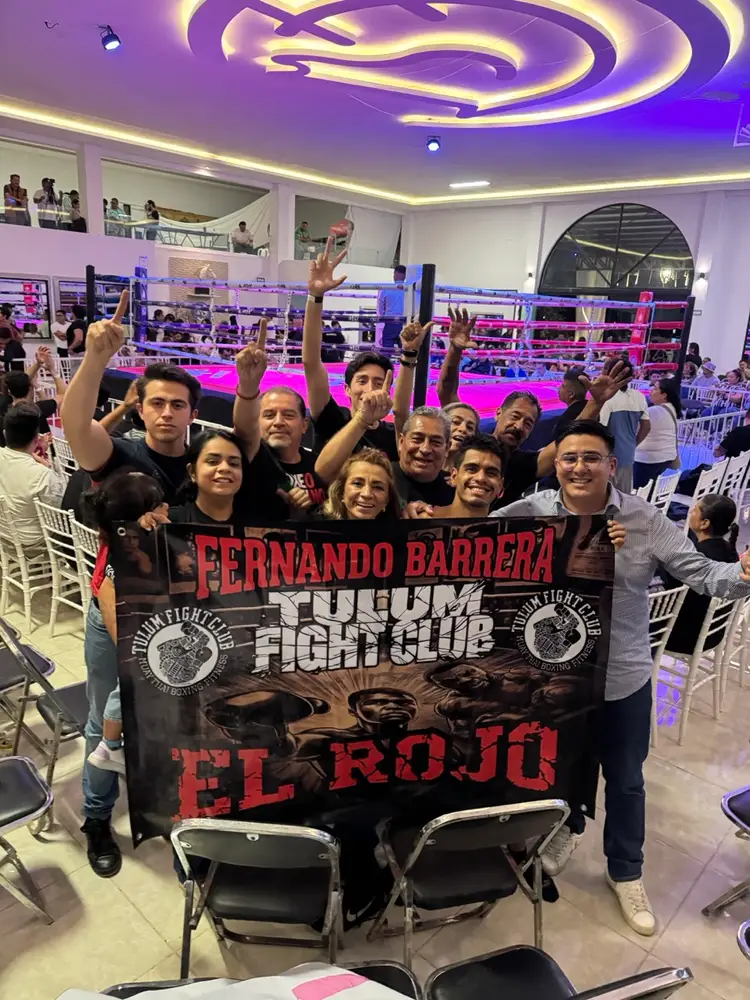 A group of people hold a banner inside an indoor boxing venue with a purple and pink neon-lit ceiling and a boxing ring visible behind them A group of people hold a banner inside an indoor boxing venue with a purple and pink neon-lit ceiling and a boxing ring visible behind them