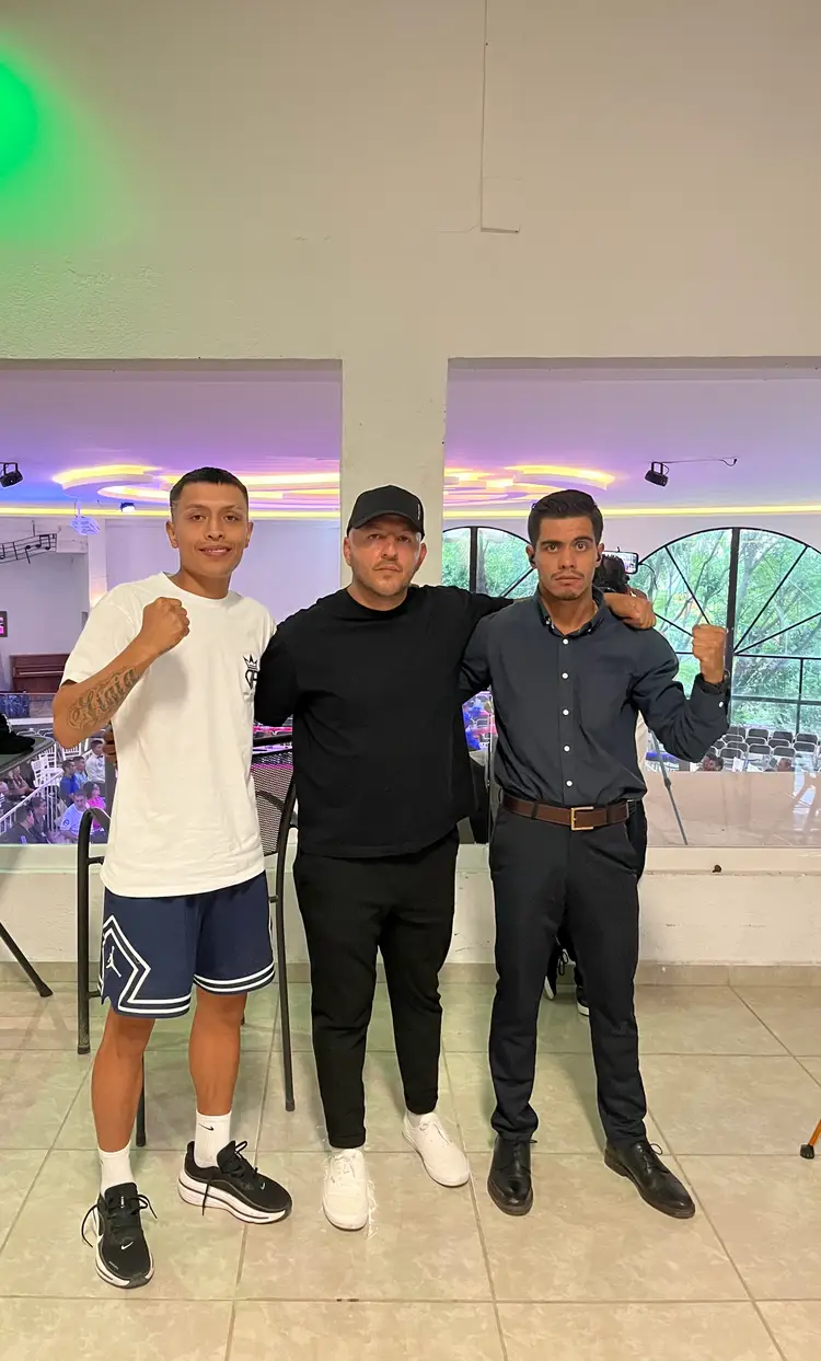 Three men pose together inside a venue with neon lighting, with two making boxing gestures toward the camera Three men pose together inside a venue with neon lighting, with two making boxing gestures toward the camera