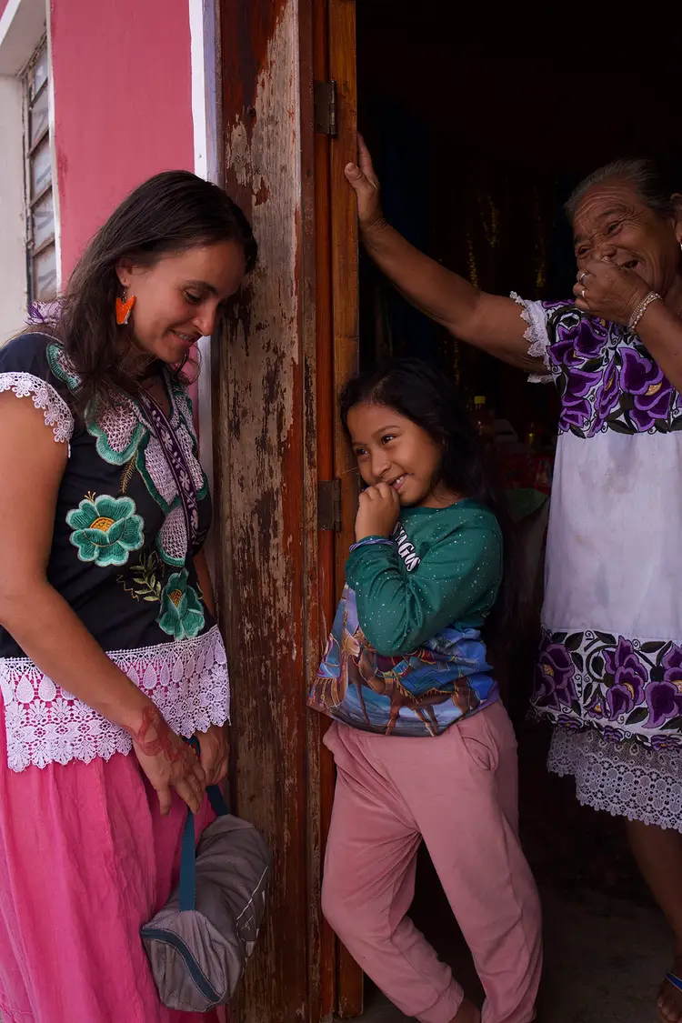 Two women and a girl stand in a doorway of a pink-walled building, smiling and interacting together in what appears to be a residential setting