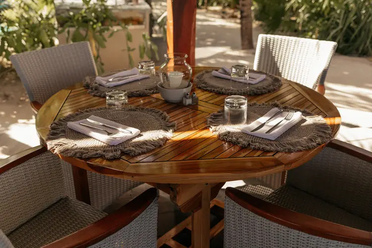 A round wooden dining table set for four with place settings, glasses, and napkins surrounded by woven chairs on an outdoor patio with greenery and a red column A round wooden dining table set for four with place settings, glasses, and napkins surrounded by woven chairs on an outdoor patio with greenery and a red column