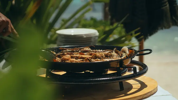 Grilled fish cooking in a paella pan over an outdoor flame, with stacked plates and tropical plants visible in the background Grilled fish cooking in a paella pan over an outdoor flame, with stacked plates and tropical plants visible in the background