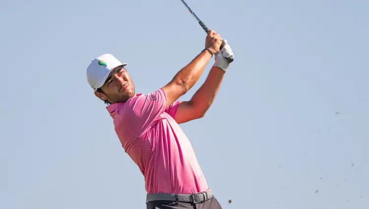 Golfer in pink shirt and white cap swings a club against a clear blue sky