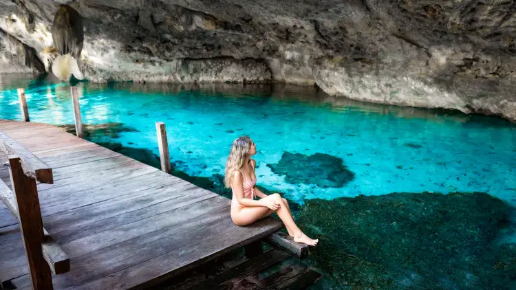 Person sitting on a wooden dock at the edge of a turquoise cenote pool beneath limestone cliffs Person sitting on a wooden dock at the edge of a turquoise cenote pool beneath limestone cliffs