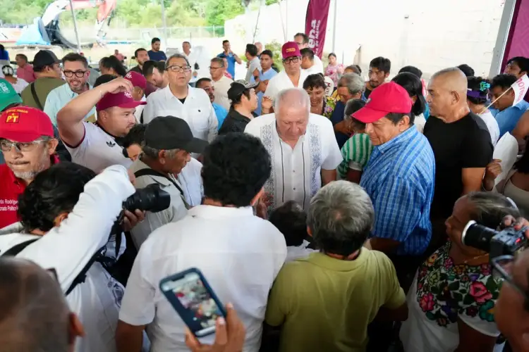 A large crowd of people gathers around a man in a white shirt at an indoor event, with some attendees holding phones to record and wearing red caps