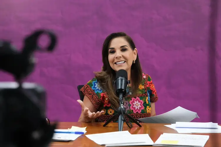 Woman speaking at a microphone during a panel or press event, seated at a table with papers and documents against a purple background
