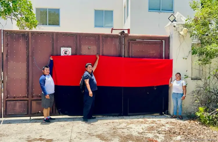 Three people hold a large red and black flag in front of a brown metal gate on a building exterior Three people hold a large red and black flag in front of a brown metal gate on a building exterior