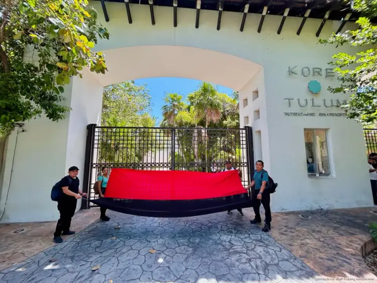 Two workers hold a large red and black banner at the entrance of a white building with arched gateway and palm trees visible beyond Two workers hold a large red and black banner at the entrance of a white building with arched gateway and palm trees visible beyond