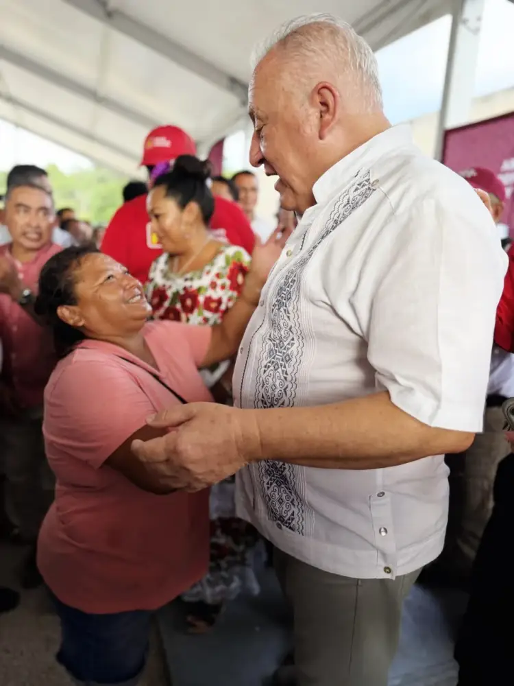Older man in white embroidered shirt shakes hands with woman in pink shirt while others in red gather under a tent structure