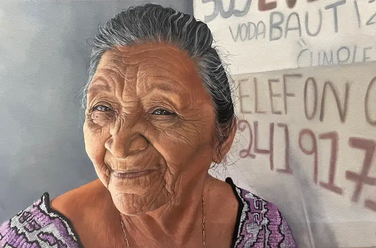 An older woman with gray hair pulled back smiles at the camera while standing in front of a wall with faded text