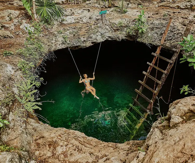 A person swings on a rope over emerald-green water inside a large underground cenote surrounded by limestone walls and wooden ladder access A person swings on a rope over emerald-green water inside a large underground cenote surrounded by limestone walls and wooden ladder access