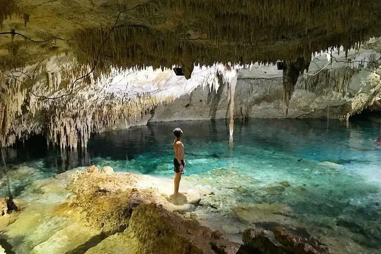 Person in swimwear stands on rocks inside a cave with turquoise water and stalactites Person in swimwear stands on rocks inside a cave with turquoise water and stalactites