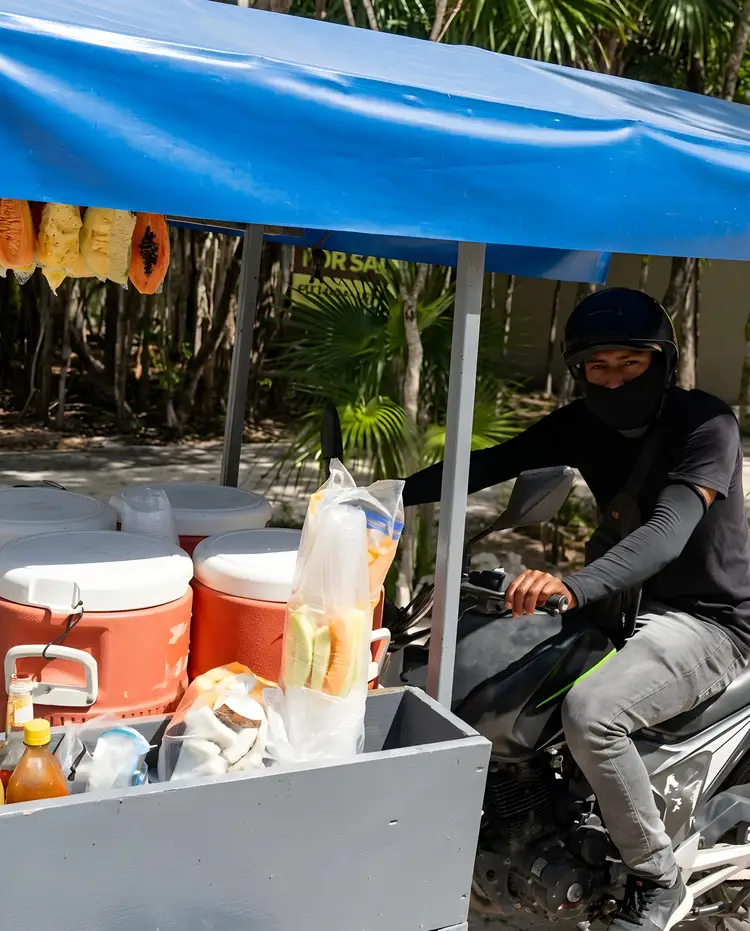 Person on a scooter operates a food cart with coolers and supplies under a blue canopy, surrounded by tropical vegetation