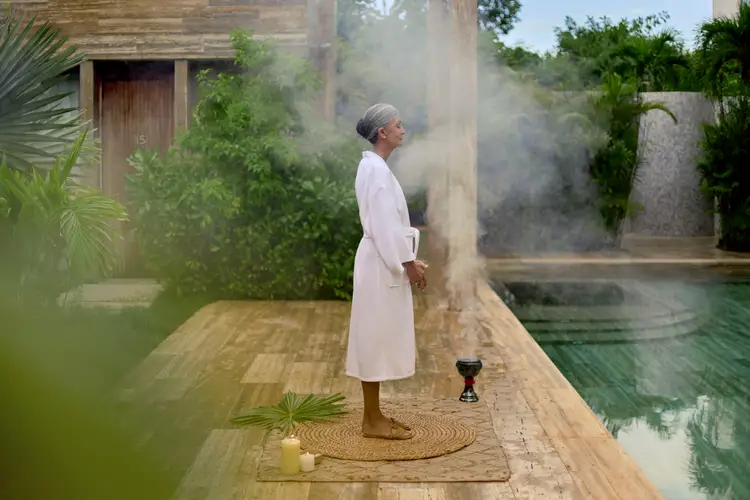 Persona con bata blanca junto a una alberca, entre humo de incienso, en el patio de un spa o resort Persona con bata blanca junto a una alberca, entre humo de incienso, en el patio de un spa o resort
