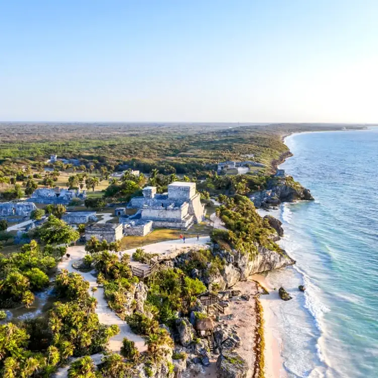 Aerial view of a white stone structure on a cliff overlooking a sandy beach and ocean, surrounded by forested landscape and coastal terrain